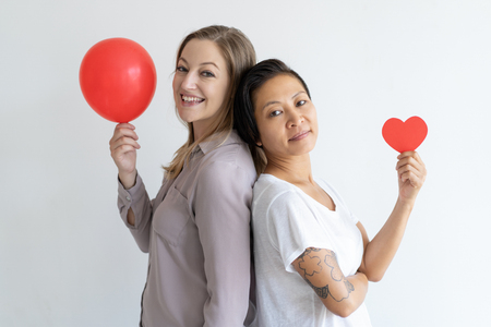 Women standing back to back with red balloon and paper heart. Multiethnic women posing at camera. Lesbian couple and Valentines day concept. Isolated side view on white background.の写真素材