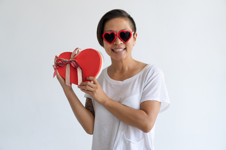 Smiling Asian woman with heart shaped gift box and party glasses. Lady wearing t-shirt and looking at camera. Saint Valentines Day concept. Isolated front view on white background.の写真素材