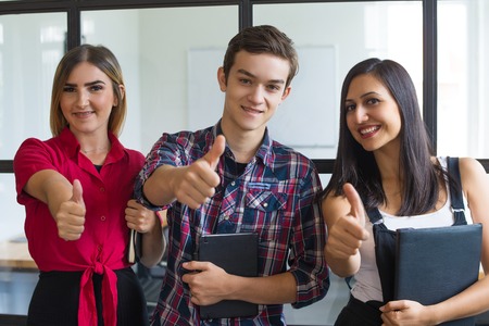 Portrait of successful young students showing thumbs up. Group of two Caucasian girls and boy...