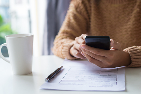 Close-up of business lady using gadget while examining document. Unrecognizable woman in warm sweater sitting at table and checking message. Technology conceptの写真素材