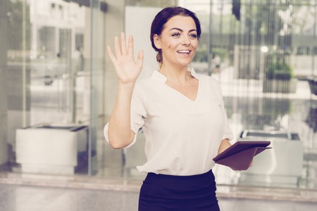 Portrait of happy mid adult secretary waving hand to colleague. Caucasian businesswoman with digital tablet greeting somebody in office. Business greeting conceptの写真素材