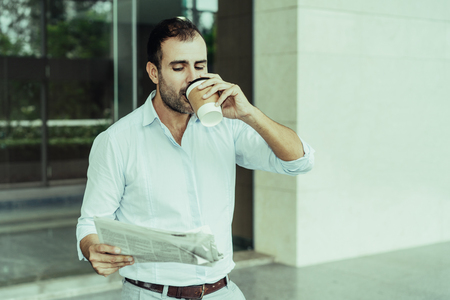 Joyful business expert enjoying coffee on his way to work. Middle aged Latin drinking takeaway coffee and reading newspaper, office building exterior in background. Morning routine conceptの写真素材