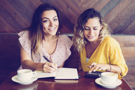 Pretty women drinking coffee and using calculator in cafe. Ladies sitting at table. Women friendship and work concept. Front view.の写真素材