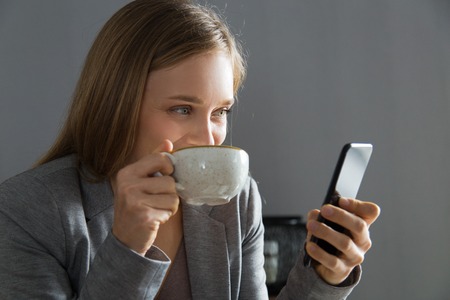 Cheerful office worker enjoying coffee break and online chat. Young woman in jacket drinking tea and...