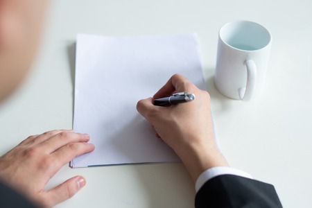 Closeup of man writing on empty sheet of paper. Business man sitting and working at office desk with cup. Workplace and document concept. Cropped top view.の写真素材