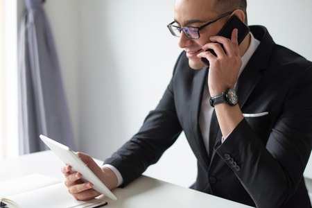 Smiling businessman using tablet and calling on smartphone. Business man sitting and working at office desk. Technologies in business concept.の写真素材