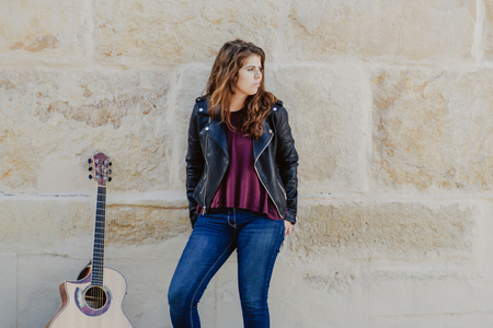 Pensive attractive female guitarist in black leather jacket standing against stoned wall and looking...