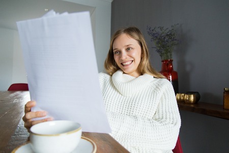 Cheerful lady in white sweater examining papers at home. Positive young lady in white sweater sitting at table and enjoying work. Paperwork conceptの写真素材