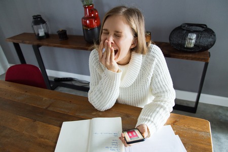 Tired young woman covering mouth while yawning. Pretty woman sitting at table and using gadget while...