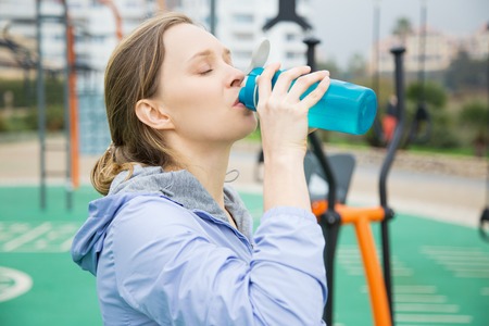 Tired fit girl feeling thirsty during physical exercises. Young woman in sports hoodie drinking...