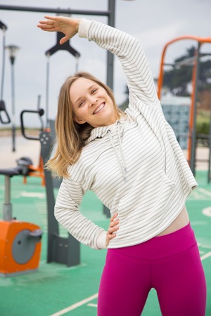 Smiling woman stretching body on sports ground outdoors. Attractive young lady standing, doing side bends and training with sports equipment in background. Street workout concept. Front view.の写真素材