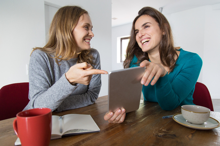 Two female friends discussing funny news. Women in casual using tablet and laughing over cup of coffee. Communication and fun conceptの写真素材