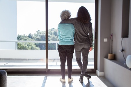 Girl visiting grandma. Back view of young and elderly women standing and looking out window. Family and taking care conceptの写真素材