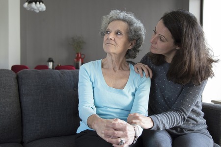 Woman talking to senior mother who is turning away. Mother and daughter talking and sitting on couch with home interior in background. Misunderstanding between mother and daughter concept.の写真素材