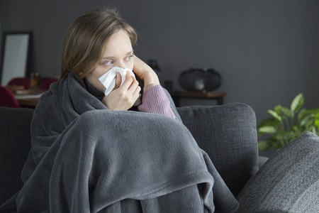 Bored sick young Caucasian woman covered with grey blanket sitting on grey sofa in living room,...