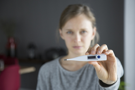 Focused close-up image of thermometer held by sick young Caucasian woman in grey sweater standing in living room, looking at camera. Illness, pain conceptの写真素材