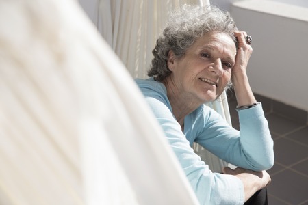 Portrait of smiling senior woman sitting in hammock on balcony. Happy retired woman posing at home. Happy retirement conceptの写真素材