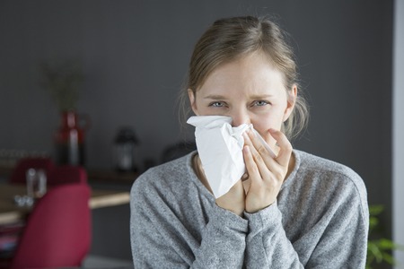 Unhappy sick young Caucasian woman in grey sweater sitting in living room, blowing her nose with napkin, looking at camera. Illness, pain conceptの写真素材