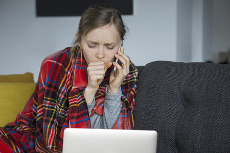 Sick young fair-haired Caucasian woman covered with plaid sitting on grey sofa in living room, coughing, working on laptop. Illness, work conceptの写真素材