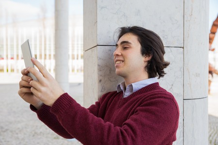 Smiling teen guy taking selfie photo on smartphone outdoors. Handsome young man wearing casual...