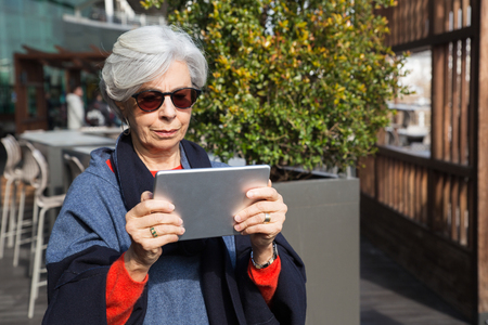 Focused senior lady consulting electronic map. Grey haired mature woman in sunglasses and overcoat standing outdoors and staring at tablet screen. Tablet using conceptの写真素材