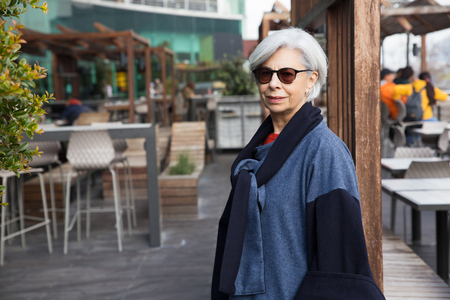 Positive senior tourist walking around city. Grey haired mature woman in sunglasses and overcoat smiling at camera, street cafes in background. Walking outdoors conceptの写真素材