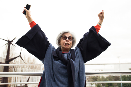 Excited senior lady with smartphone celebrating success. Grey haired mature woman in sunglasses and overcoat throwing hands in air and enjoying win. Success gesture conceptの写真素材