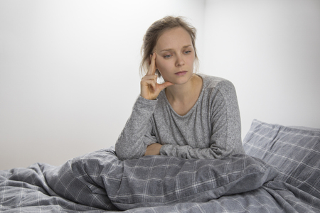 Depressed sick young Caucasian woman in grey homewear sitting on bed with pillow on her knees, looking aside. Illness, recovery conceptの写真素材