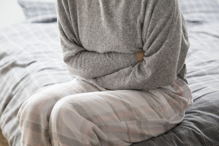 Sick woman in grey homewear sitting on bed, keeping hands on stomach, suffering from pain. Illness, stomach ache conceptの写真素材