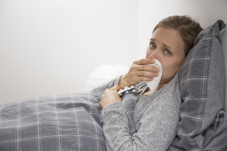 Sick young Caucasian woman in grey homewear lying in bed on her back, turning head to camera,...