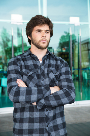 Pensive young man standing outdoors with his arms crossed. Handsome guy wearing casual shirt, looking away and standing with glass wall in background. Young man portrait concept. Front view.の写真素材