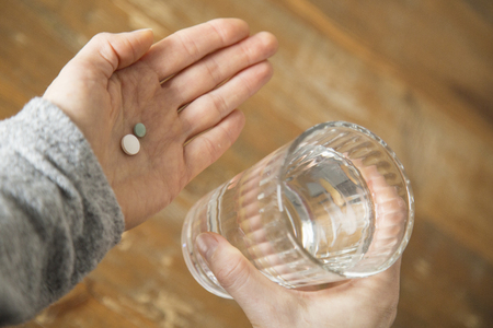 Closeup of woman holding pills on palm and glass of water. Sick woman taking medicine at home. Disease and medical treatment concept. Cropped view.の写真素材