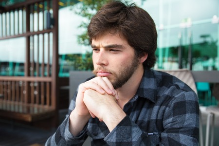 Upset young man thinking over bad news or problems. Frowning guy in casual shirt sitting in street cafe, leaning chin on clasped hands and thinking hard. Bad news conceptの写真素材