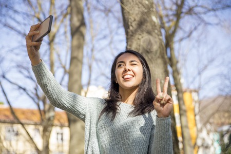 Carefree woman grimacing and taking selfie photo outdoors.  Smiling pretty young lady posing, showing victory sign and using smartphone with trees in background. Selfie photograph concept.の写真素材