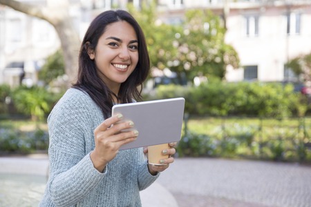 Happy woman using tablet computer and holding plastic cup outdoors. Pretty lady looking at camera and standing with green plants and buildings in background. Urban lifestyle and technology concept.の写真素材