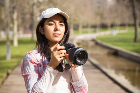 Focused young photojournalist working outdoors. Young woman wearing cap holding photo camera at face and looking away. Occupation conceptの写真素材