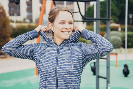 Smiling young woman putting hoodie hood on sports ground. Attractive sporty lady listening to music and looking away with sports equipment in background. Outdoor workout concept. Front view.の写真素材