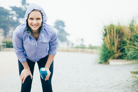Cheerful jogger catching breath. Young woman in sports hoodie and tights leaning forward, holding water bottle and smiling. Jogging or stress relief conceptの写真素材