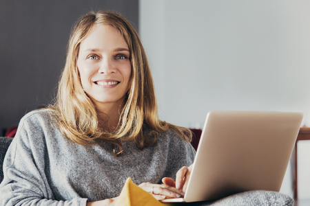 Happy freelancer at work. Positive young woman sitting on couch, using laptop and smiling at camera. Remote work conceptの写真素材