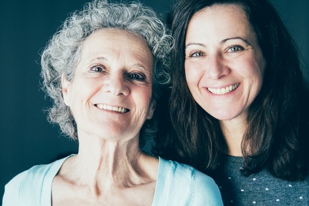 Smiling senior woman and her daughter posing at camera. Mother and daughter with grey wall in background. Family portrait concept. Front view.の写真素材