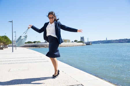 Cheerful business lady celebrating success. Happy carefree young woman in formal suit and sunglasses dancing on promenade by river. Good news conceptの写真素材
