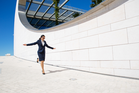 Ecstatic business lady jumping outdoors. Happy joyful young woman in formal suit running with spread hands pass modern city greenhouse. Euphoria conceptの写真素材