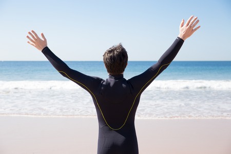 Back view of man wearing wetsuit and raising arms on sunny beach. Guy standing with sea in background. Tourism and freedom concept. Back view.の写真素材