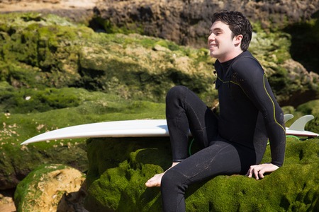Happy man sitting on mossy rocks with surfboard. Handsome young guy wearing wetsuit. Surfboarding and tourism concept.の写真素材