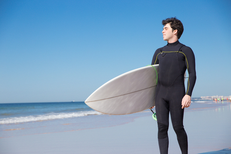 Smiling handsome young man holding surfboard on sunny beach. Handsome guy wearing wetsuit and standing. Surfboarding concept.の写真素材