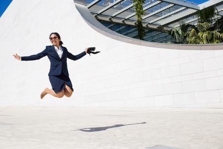 Joyful excited businesswoman celebrating success. Positive young woman in formal suit holding shoes and jumping high outdoors. Business success conceptの写真素材