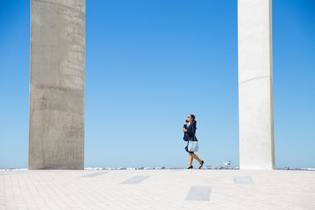Stylish business lady walking along promenade by sea and talking on phone. Wide shot of young woman...