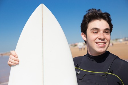 Smiling young man with surfboard on sunny beach. Handsome guy wearing wetsuit and standing. Surfer portrait concept. Closeup front view.の写真素材