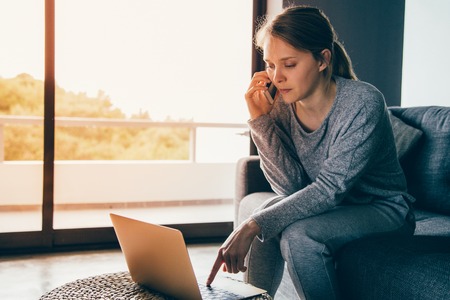 Sick young Caucasian woman in grey homewear sitting on grey sofa in living room, talking on phone, working on laptop. Illness, work conceptの写真素材