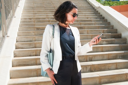 Medium shot of concentrated good looking businesswoman in white jacket with blue bag standing on stairs, looking at phone. Side view. Work, communicationの写真素材